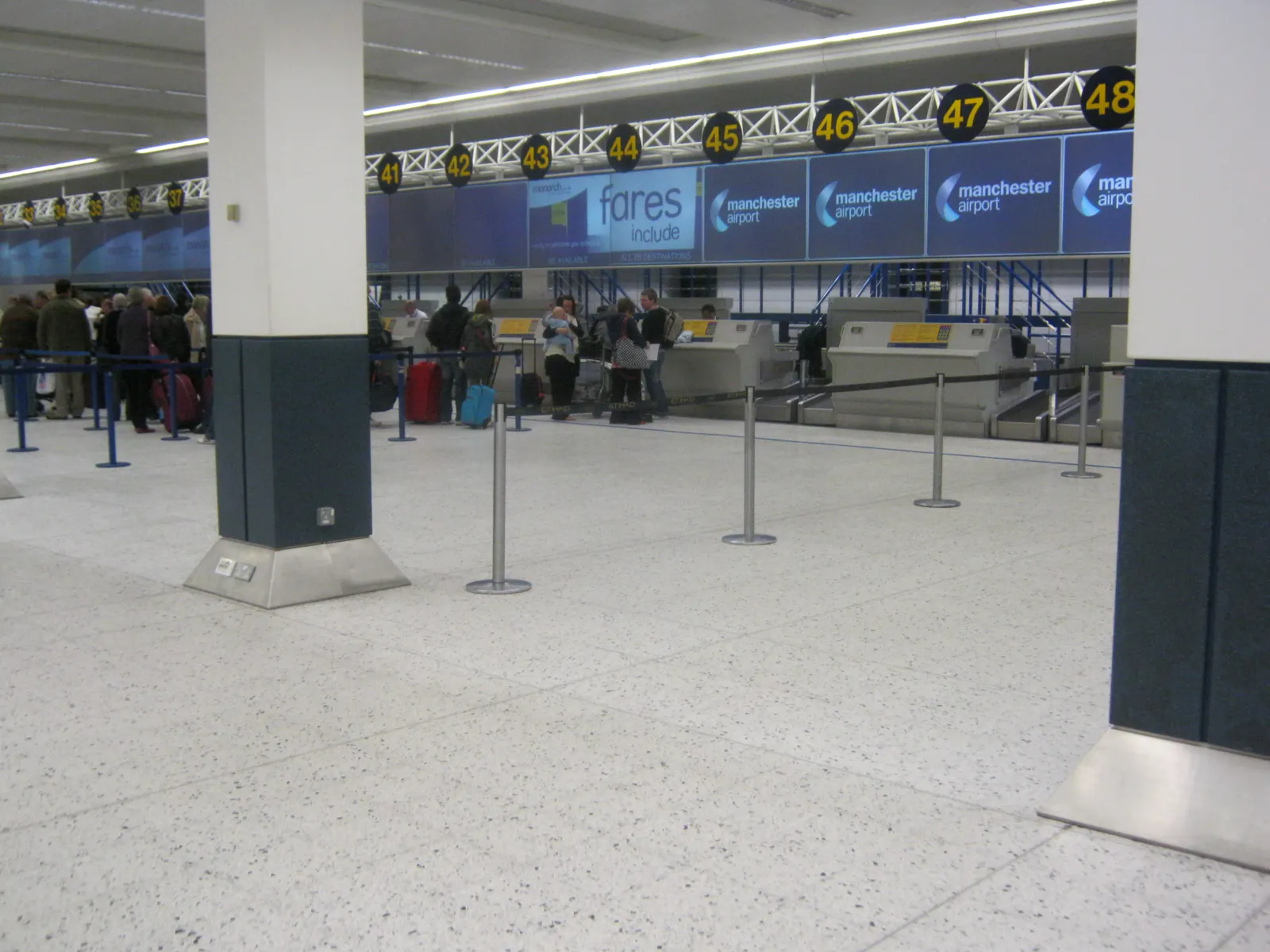Wide view of the check-in hall in Manchester Airport Terminal 1 with airline counters and travelers queuing at desks.