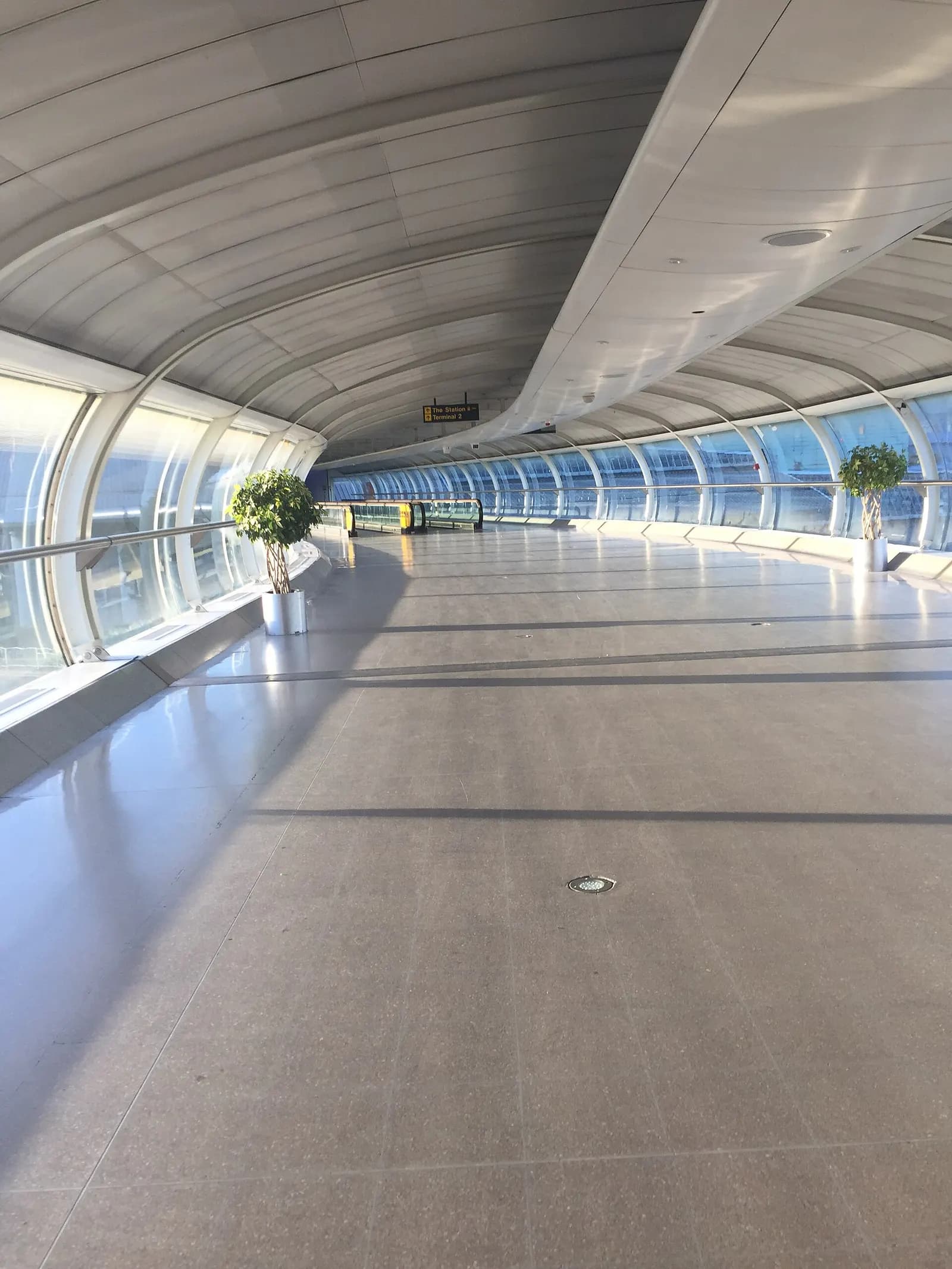 Glass-enclosed elevated skybridge walkway inside Manchester Airport connecting terminal buildings.