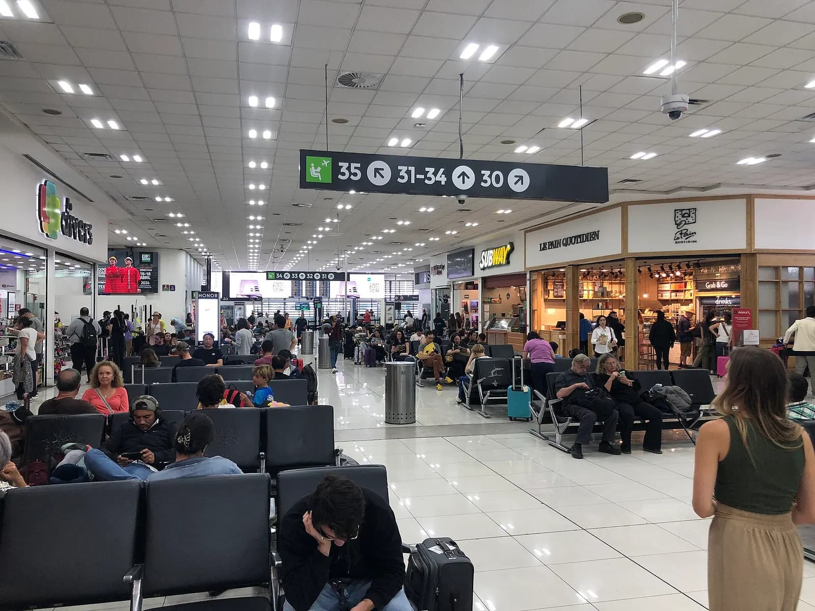 Departures gate area in Terminal 1 with seating, flight-information displays, and boarding gates along the concourse.