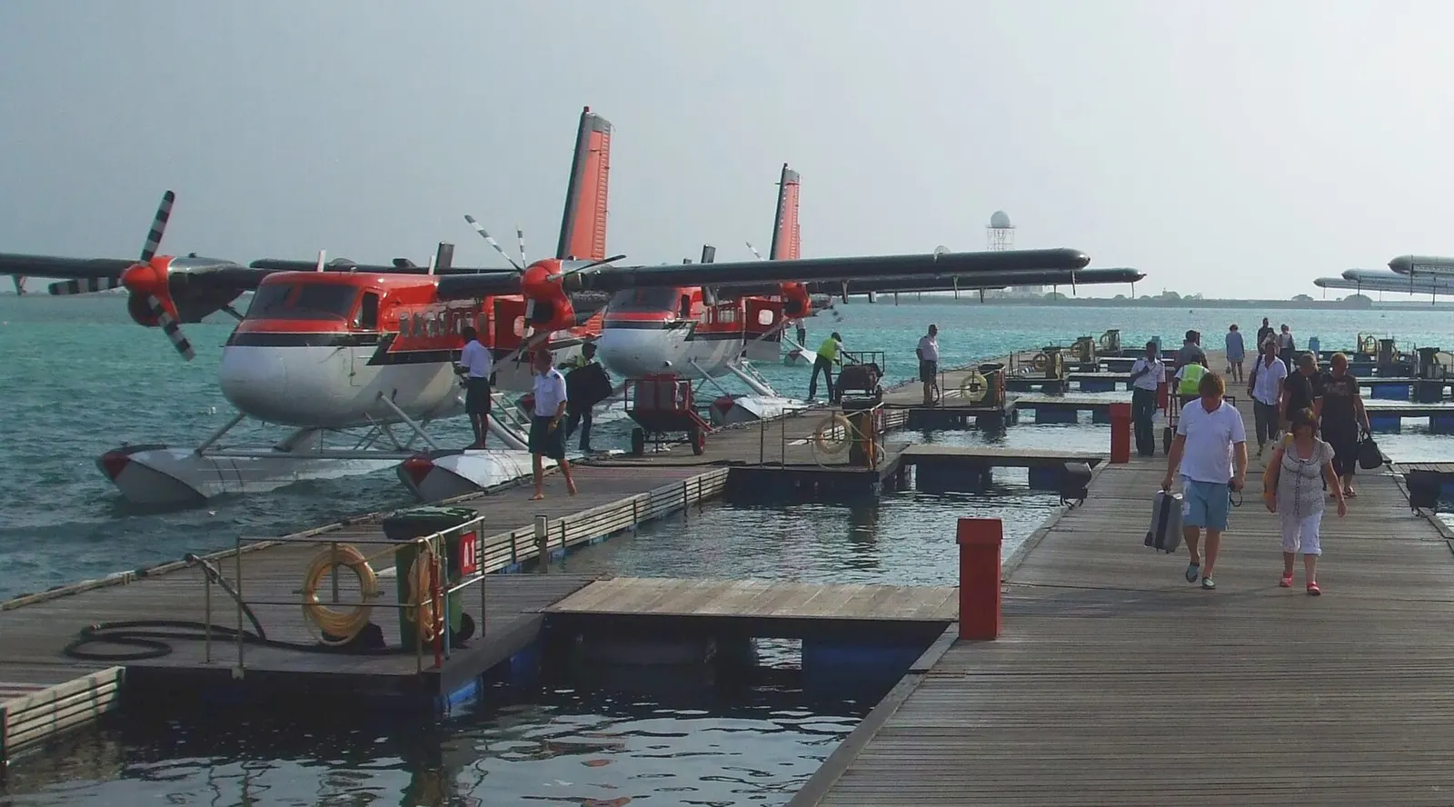 Passengers and crew on wooden seaplane docks with Trans Maldivian Airways Twin Otters at Malé