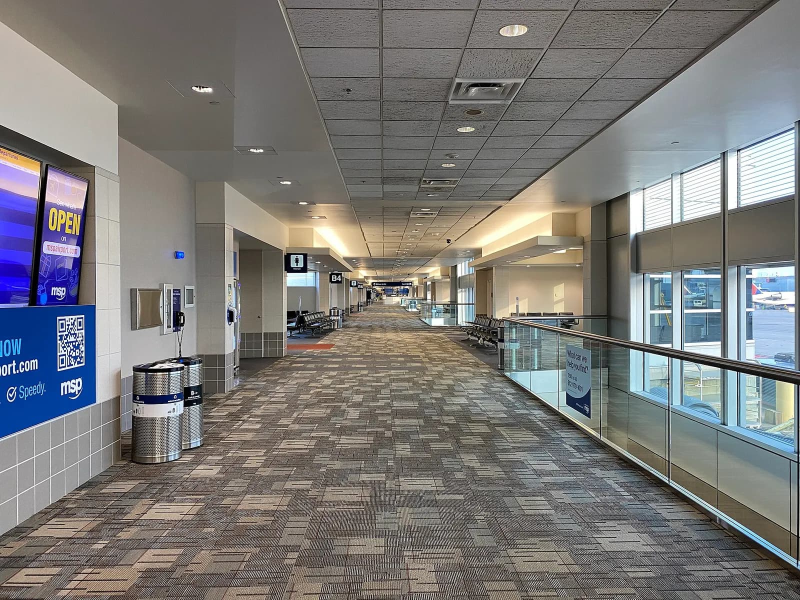 A long concourse corridor in Terminal 1 Concourse B with gate seating areas, flight information displays, and large windows overlooking the airfield.