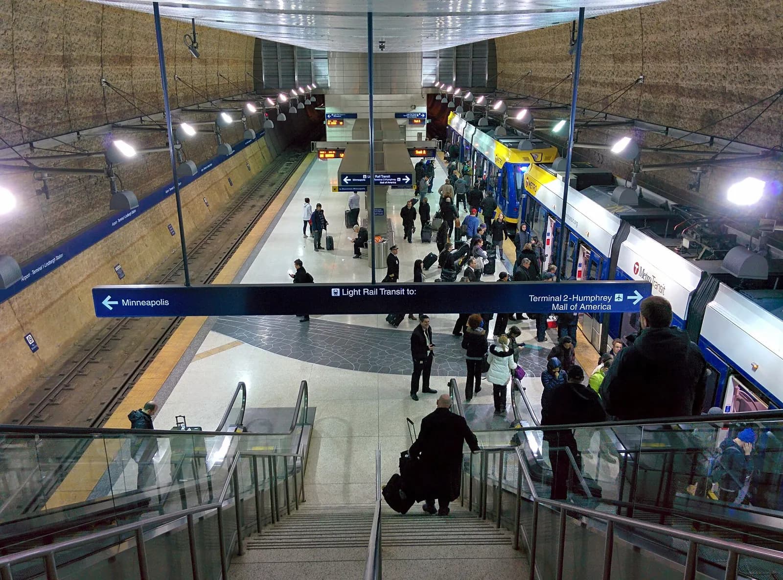 The covered METRO Blue Line light rail platform at Terminal 1 with trains, signage, and escalators leading to the airport terminal.