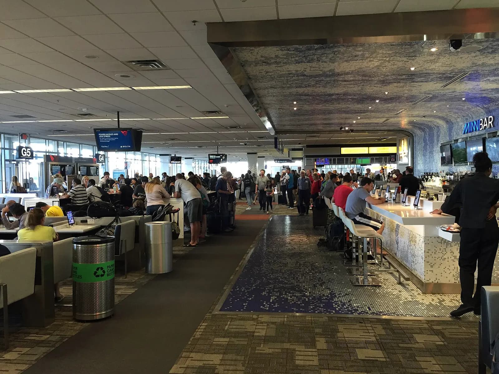 Passengers seated in curved gate-area chairs beneath high windows in the Lindbergh Terminal, with shops and walkways visible in the background.