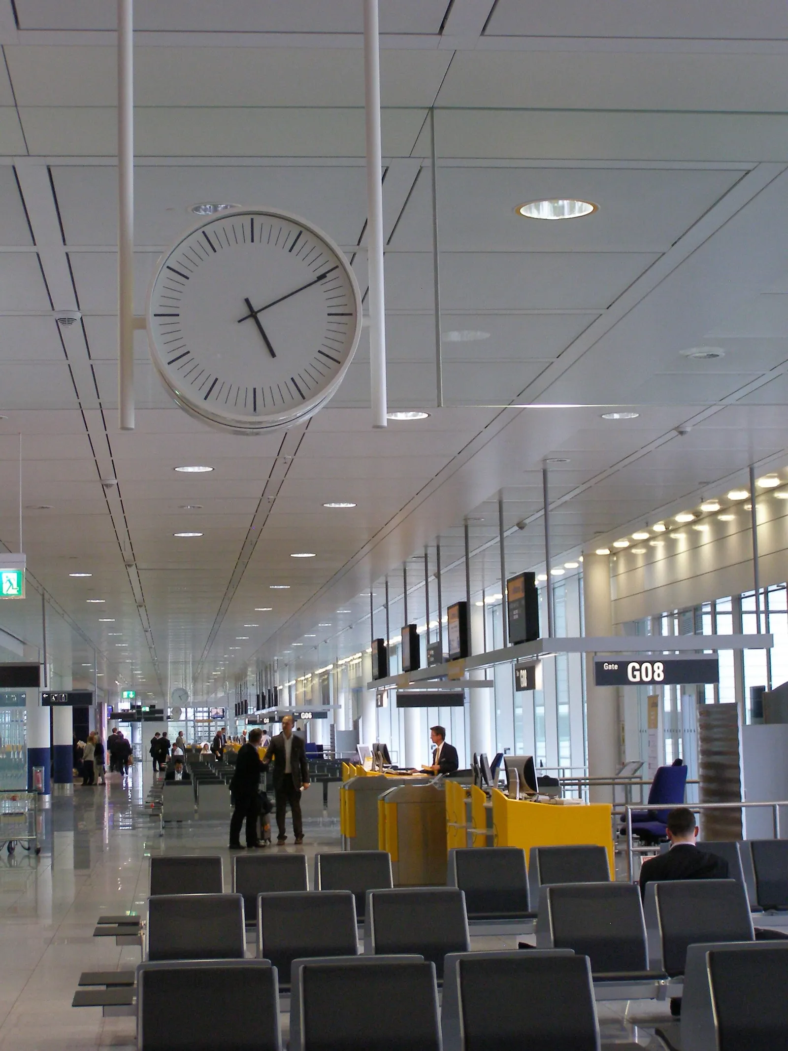 A row of departure gates with seating areas, flight information displays, and large windows overlooking the apron.