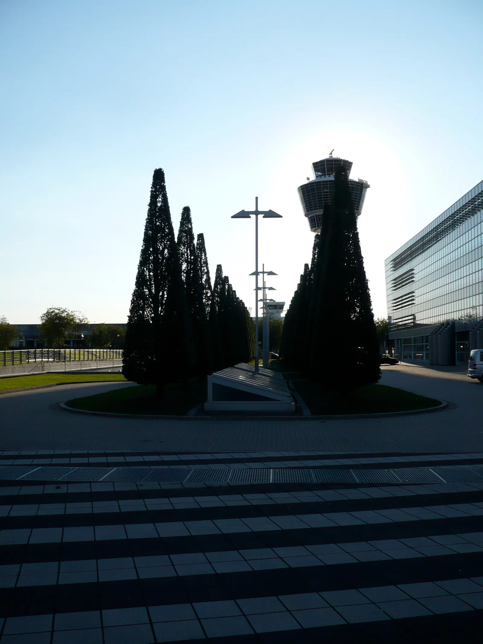 A ground-level view between parking garage P20 and the Munich Airport Center looks toward the Terminal 1 B entrance and its distinctive roofline.