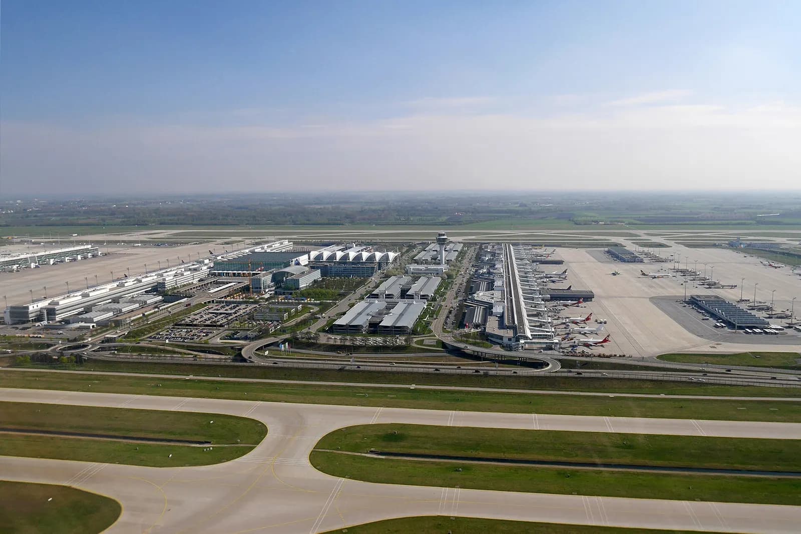 An aerial photograph shows Munich Airport's Terminal 1, Terminal 2, and the Munich Airport Center complex surrounded by runways and taxiways.