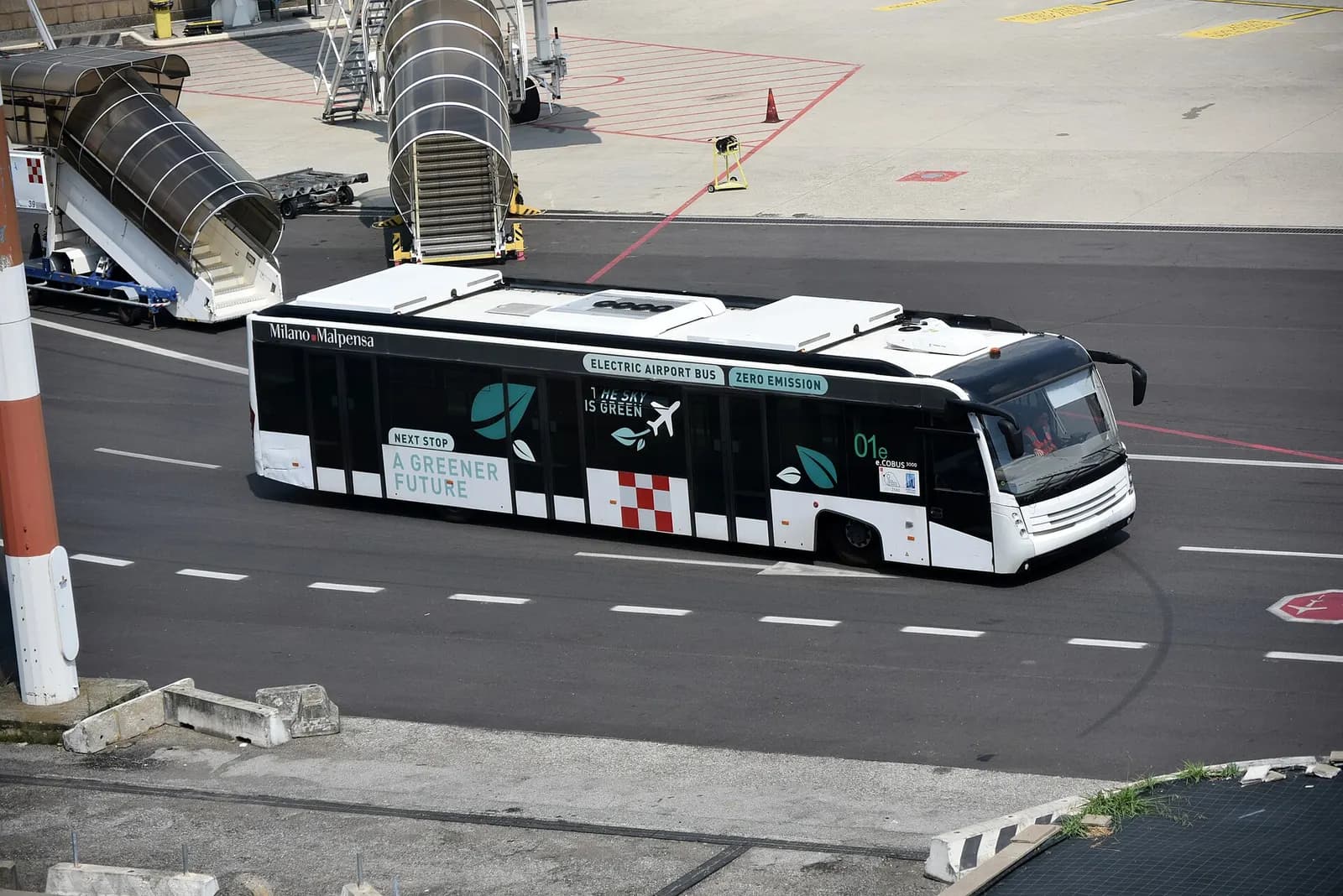 A white electric Cobus apron bus labeled for Milan Malpensa drives on the tarmac near Terminal 1.