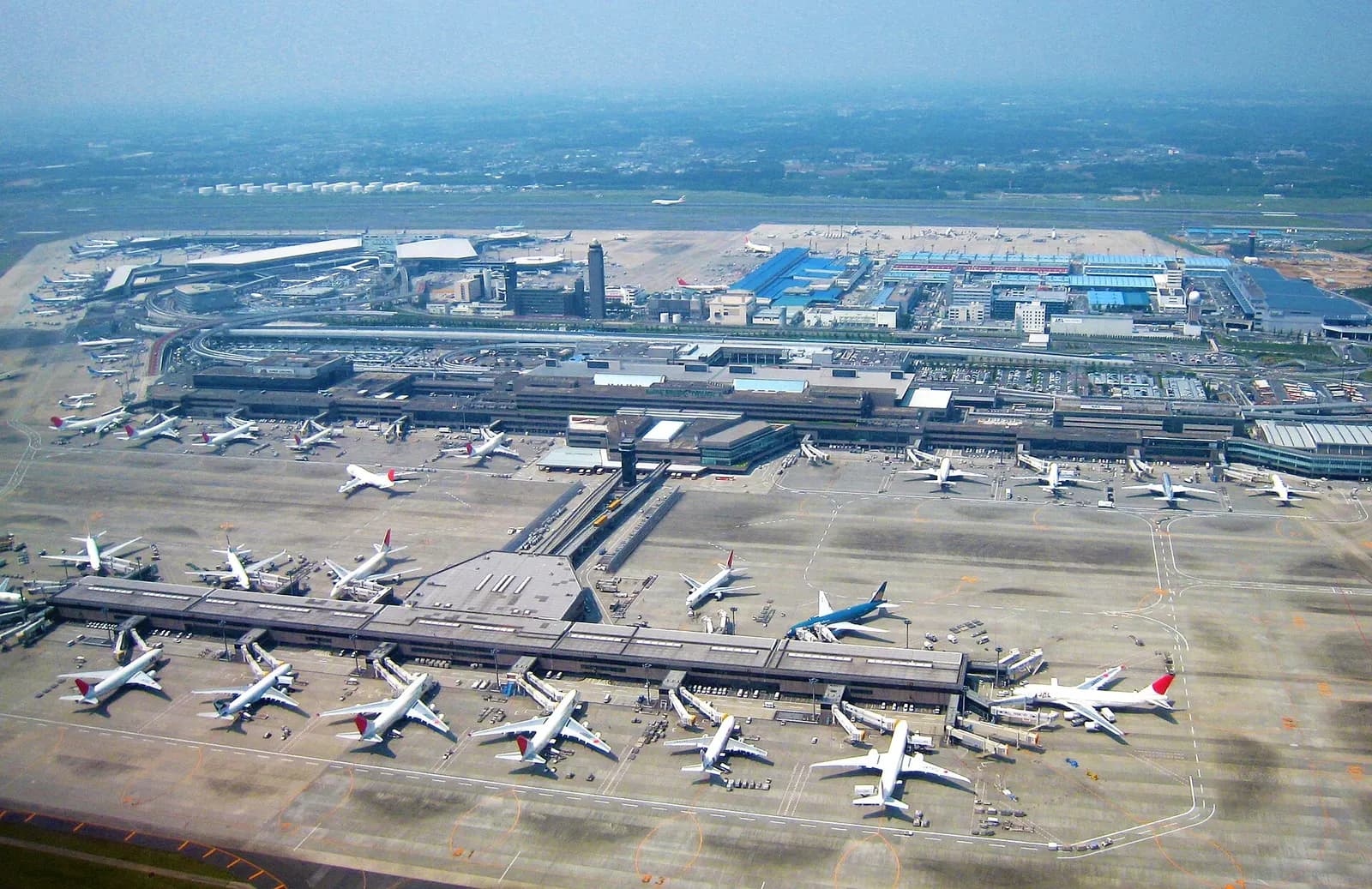 An aerial view of Narita International Airport showing multiple runways, terminal complexes, and aircraft spread across a large airfield surrounded by green countryside.