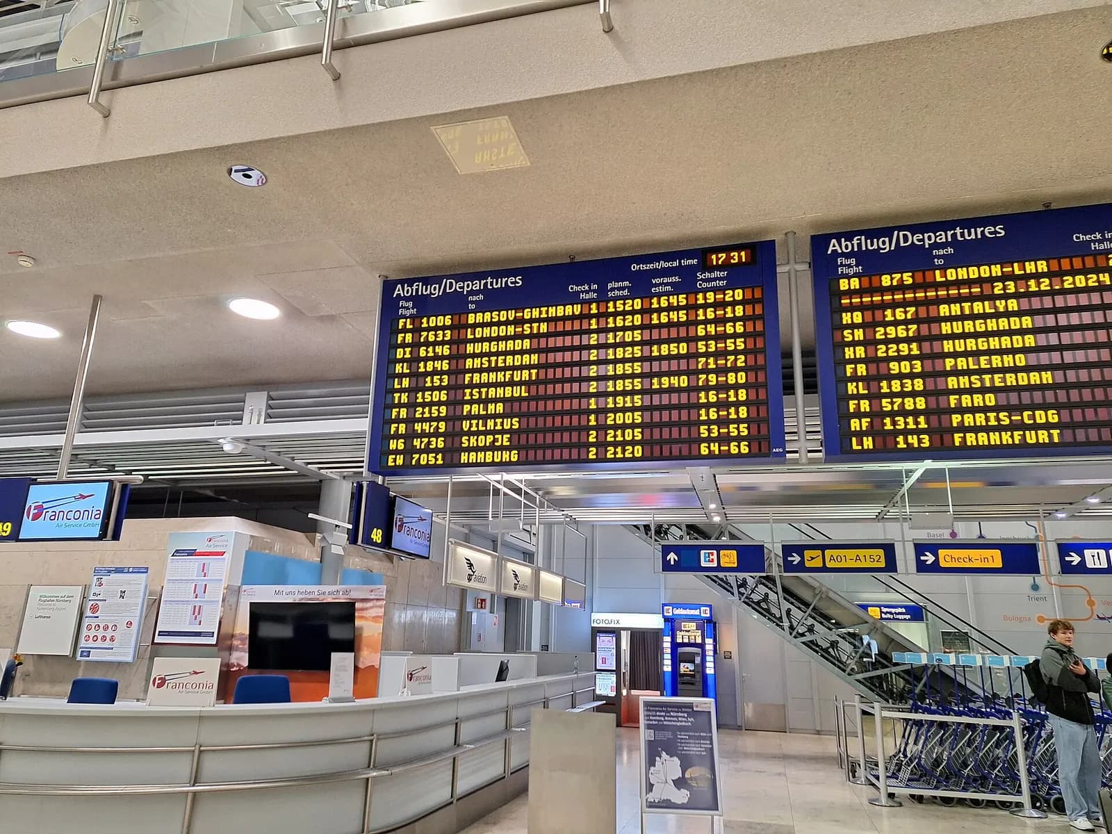 Departure information displays in the departures hall of Nuremberg Airport (Flughafen Nürnberg) on 22 December 2024. The electronic boards show various flights, destinations, scheduled and updated departure times, and check-in counters. In the foreground are the “Franconia” service desks; in the bac
