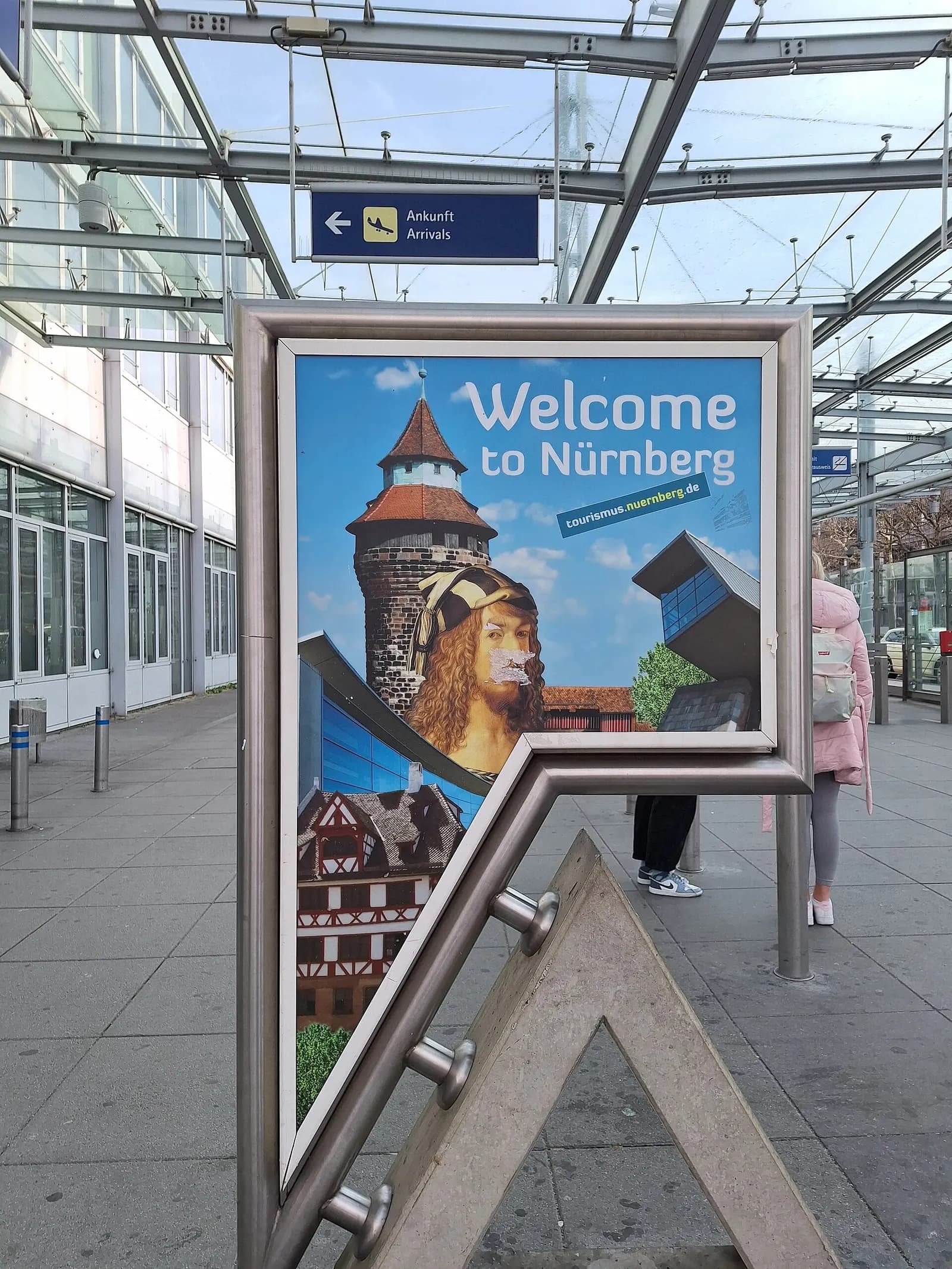 Advertising board at Nuremberg Airport displaying the message “Welcome to Nürnberg”. The welcome poster features several iconic elements of the city, including a tower of Nuremberg Castle, a portrait of Albrecht Dürer, and traditional half-timbered houses. In the background are the covered entrance 