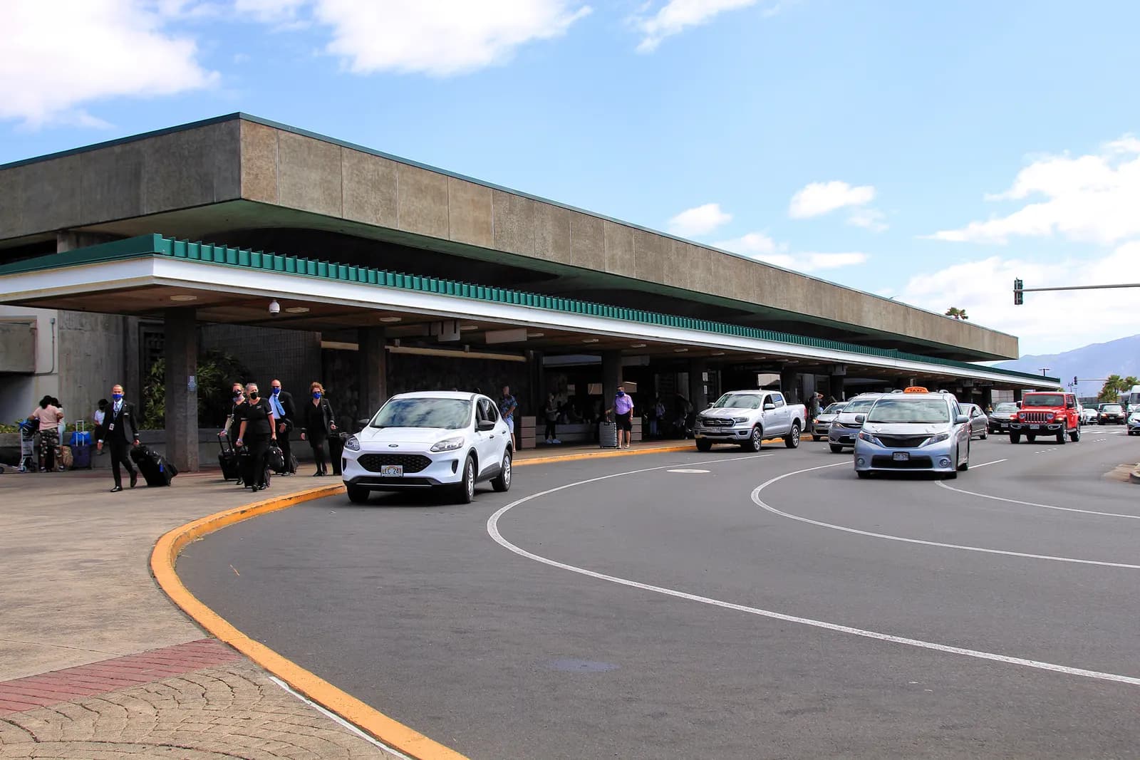 Landside curb and concrete canopy at Kahului Airport in 2021, with Hawaii plates and masked travelers on the sidewalk.