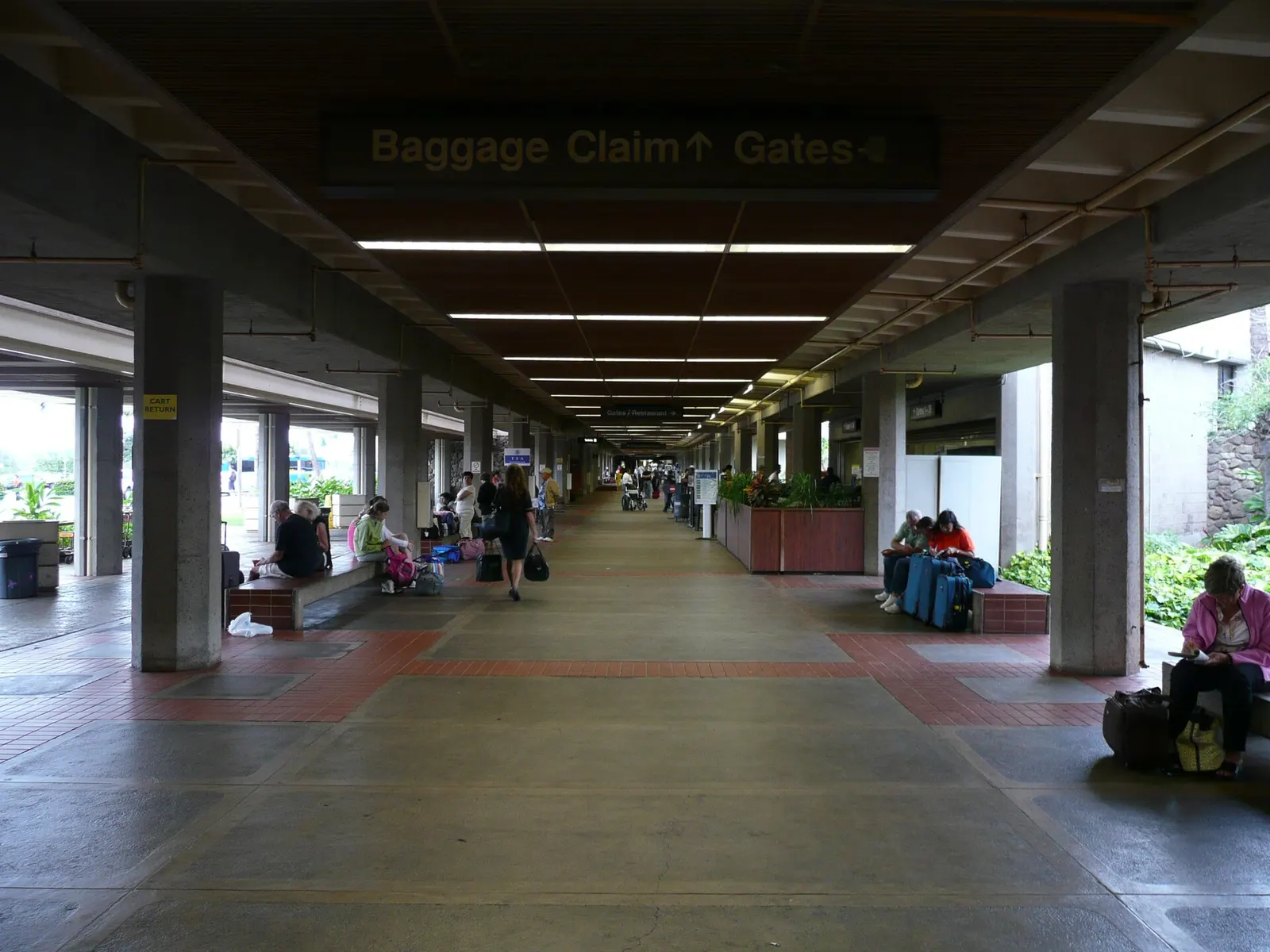 Open-air Kahului walkway with a Baggage Claim and Gates sign, lava-rock planter wall, and travelers on benches.