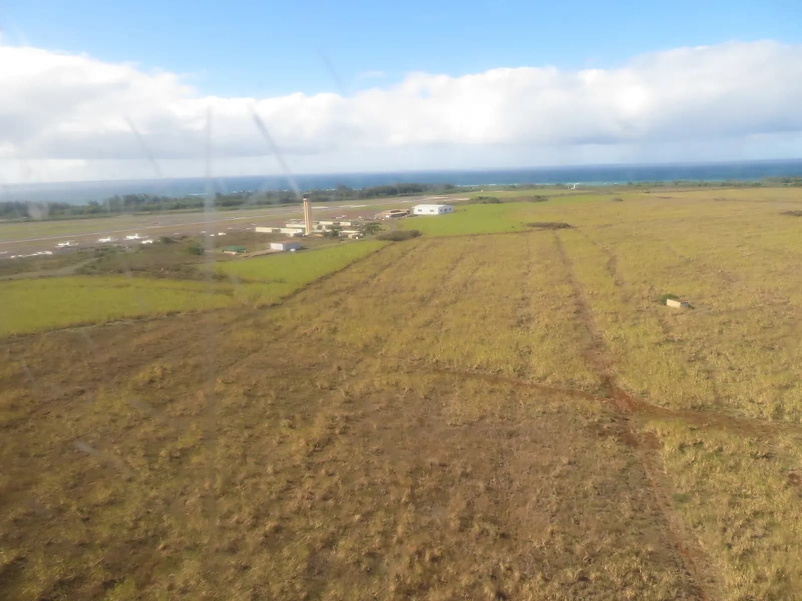 Aerial of Kahului’s 187-foot control tower, east helipad, and the shoreline runway with dry cane fields in the foreground.
