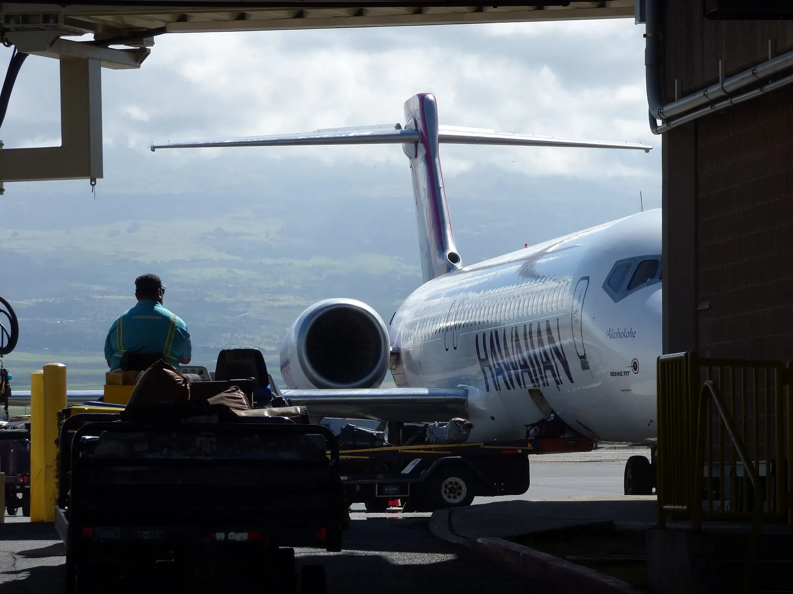 Hawaiian Airlines 717 ‘Akohekohe on a Kahului jetbridge with a baggage tug in the foreground and Haleakalā in cloud behind.