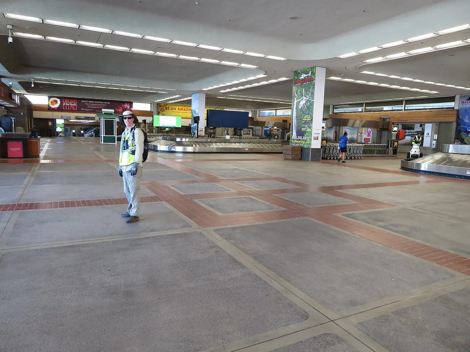 Kahului baggage-claim hall with circular carousels, luggage carts, and Maui helicopter / luau ads — nearly empty in May 2020.