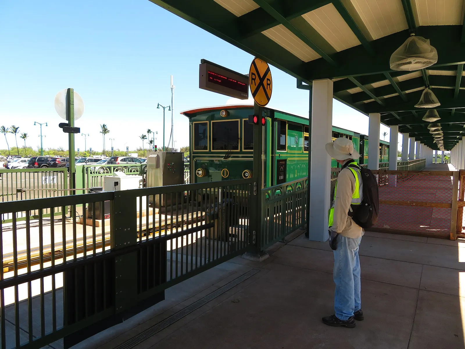 Green CONRAC tram car 1 at a covered Kahului Airport platform, with palm trees and a rental-car parking lot behind.