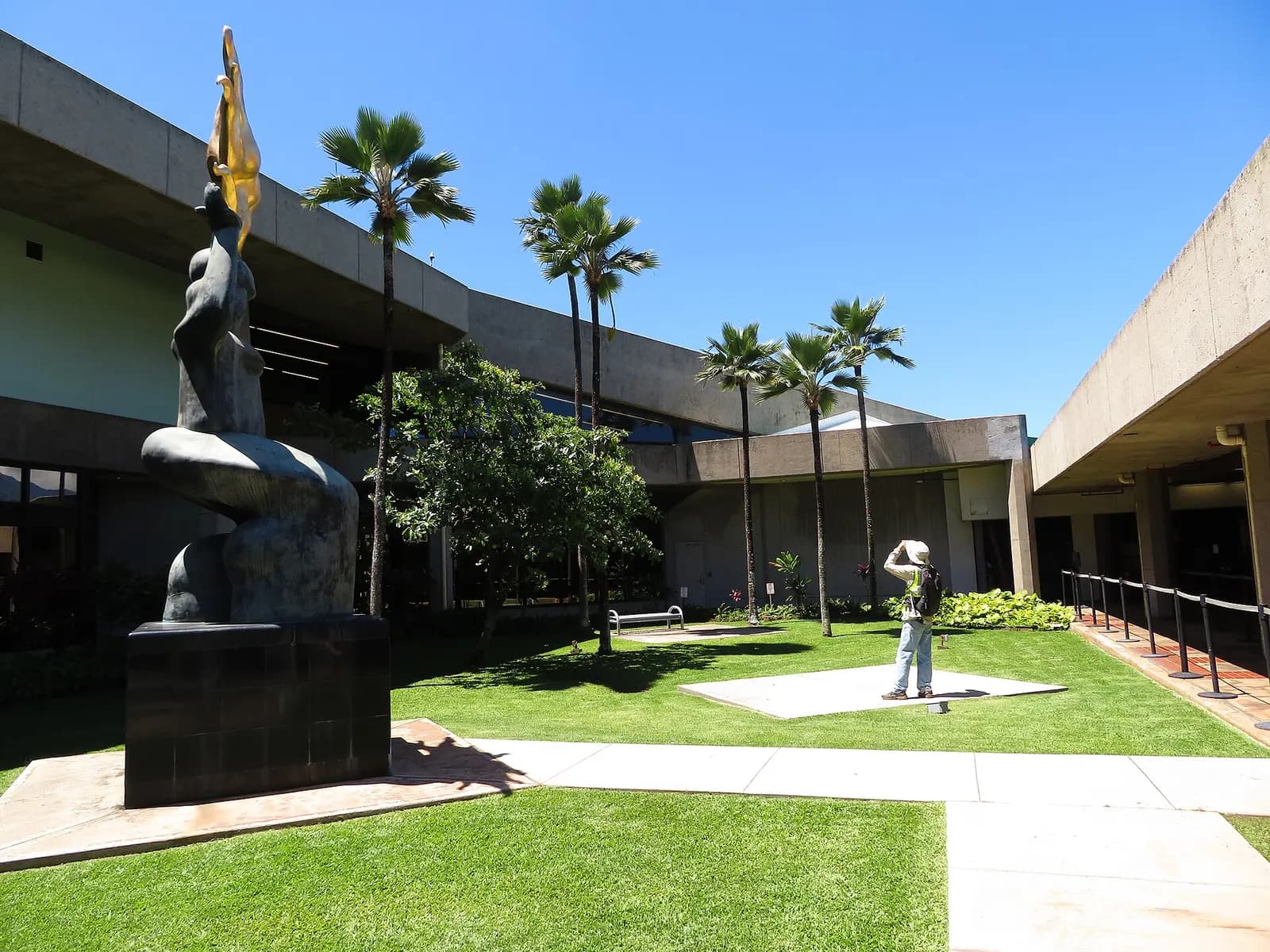 Open courtyard at Kahului Airport with the bronze Maui Releasing the Sun statue, palms, and the concrete terminal wings.