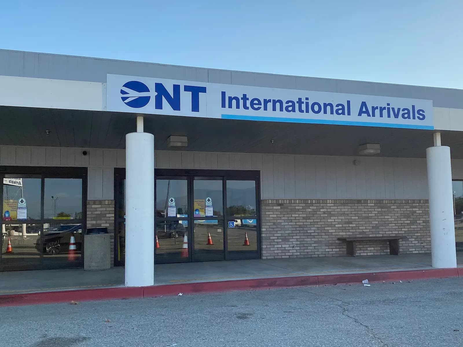 Beige-brick entrance marked ONT International Arrivals with automatic glass doors and a wooden bench.