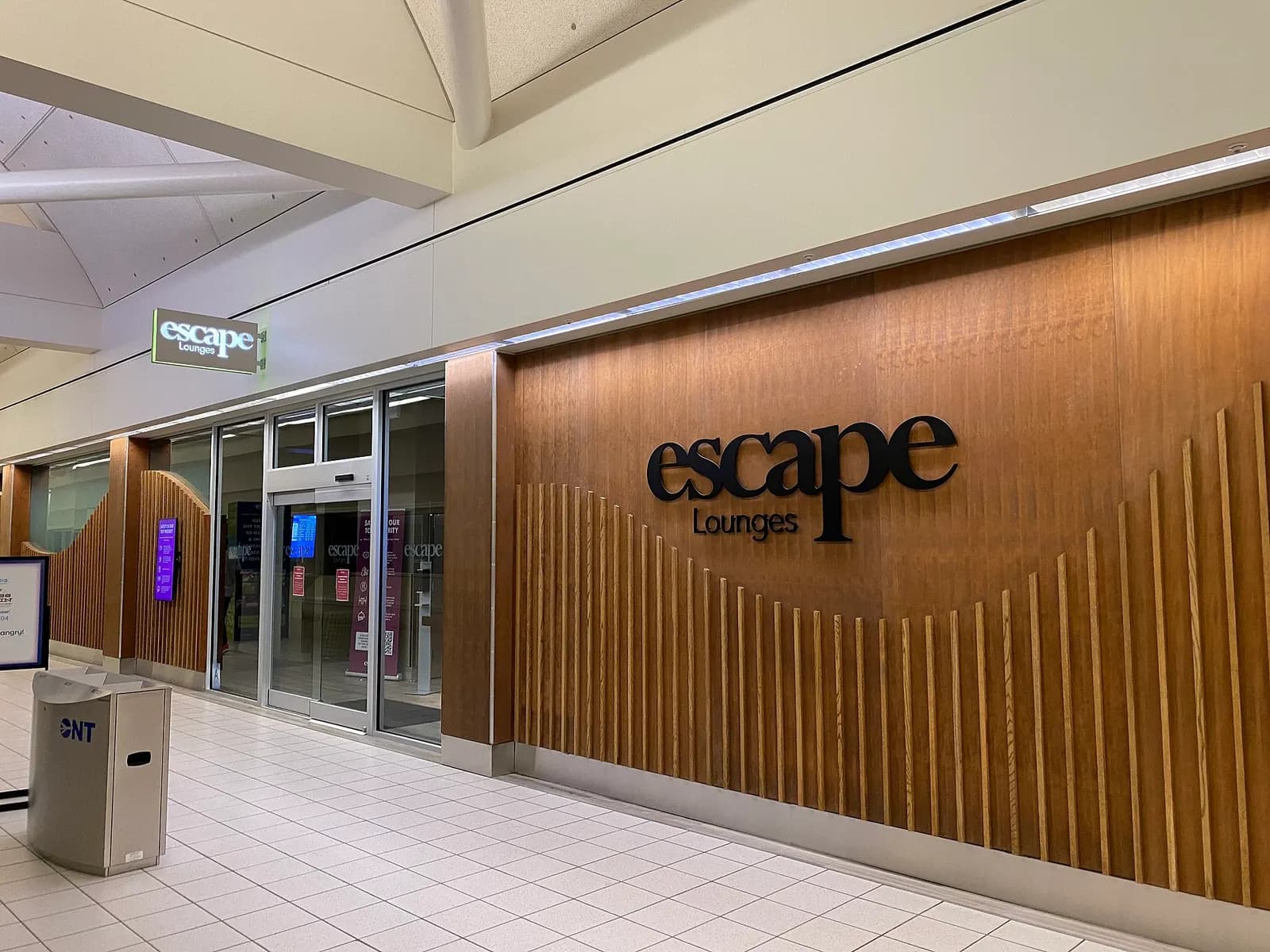Wood-paneled Escape Lounges entrance in an ONT concourse with an ONT-branded trash can in the foreground.