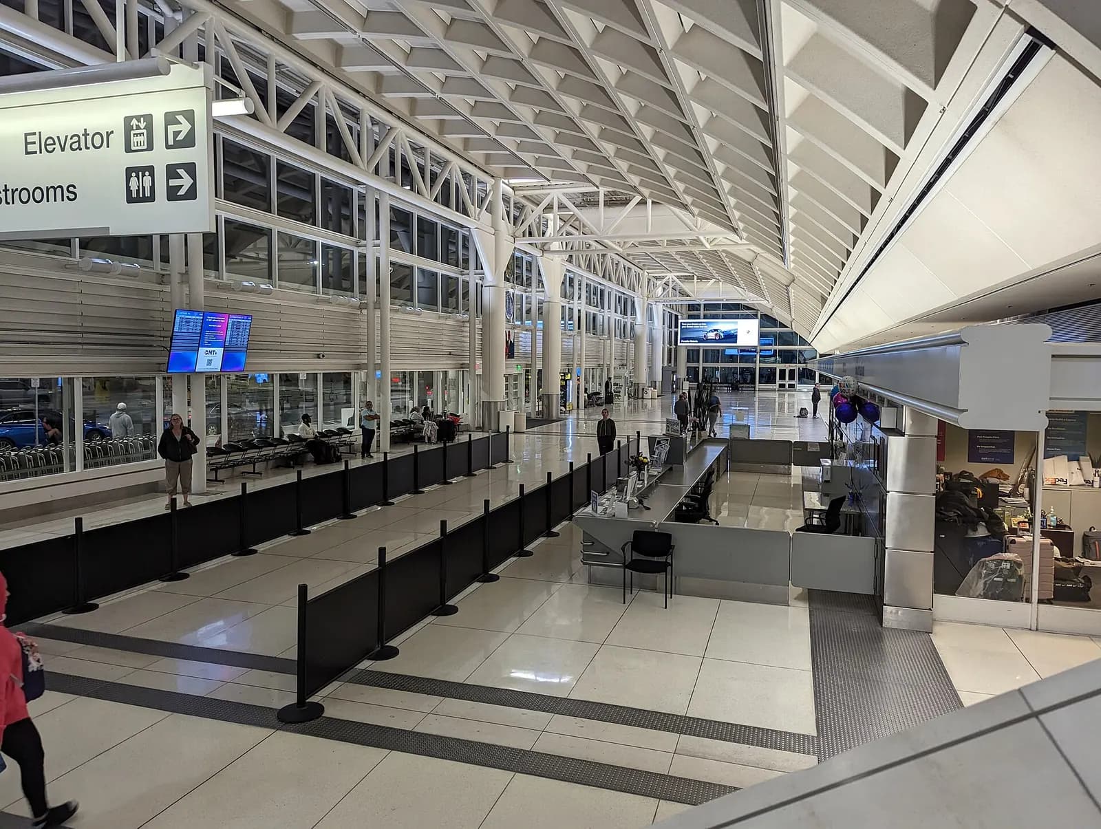Night interior of Ontario Terminal 2 with the vaulted white truss ceiling, queue stanchions, and an ONT flight-information screen.
