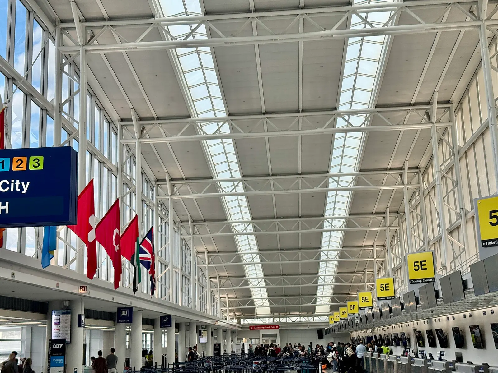 The bright, open entrance hall of Terminal 5 features tall windows, check-in kiosks, and passengers pulling luggage.