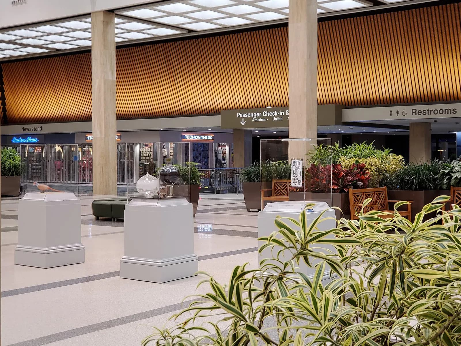Main Lobby landside with live plants, Hudson News, Tech on the Go, and wayfinding to passenger check-in and restrooms.