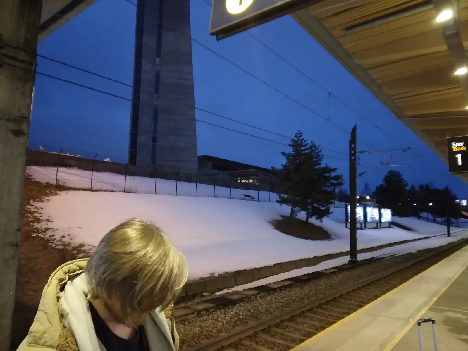 Oslo Lufthavn train station platform at dusk, with railway tracks, overhead wires, and the station structure beside the airport terminal.