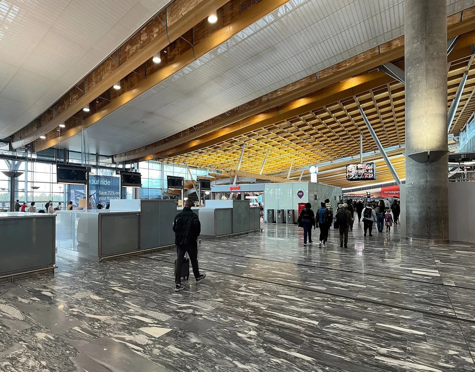The curved glass-and-wood facade of Oslo Airport Gardermoen Terminal 1 under a cloudy sky, with approach roads and landscaping in the foreground.