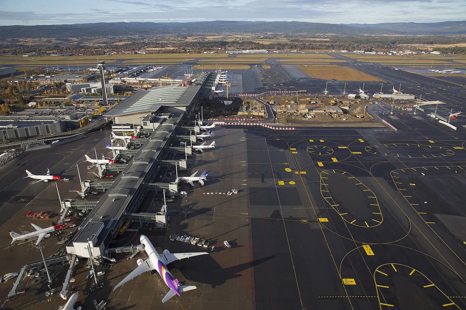 Aerial view of Oslo Airport showing parallel runways, terminal buildings, parked aircraft on the apron, and surrounding forest.