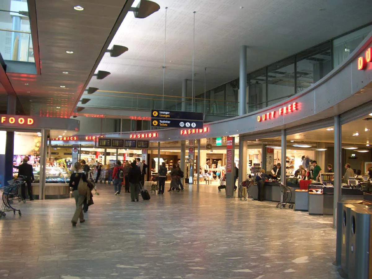 Duty-free shopping corridor inside Oslo Airport with retail displays, bright signage, and passengers browsing stores.