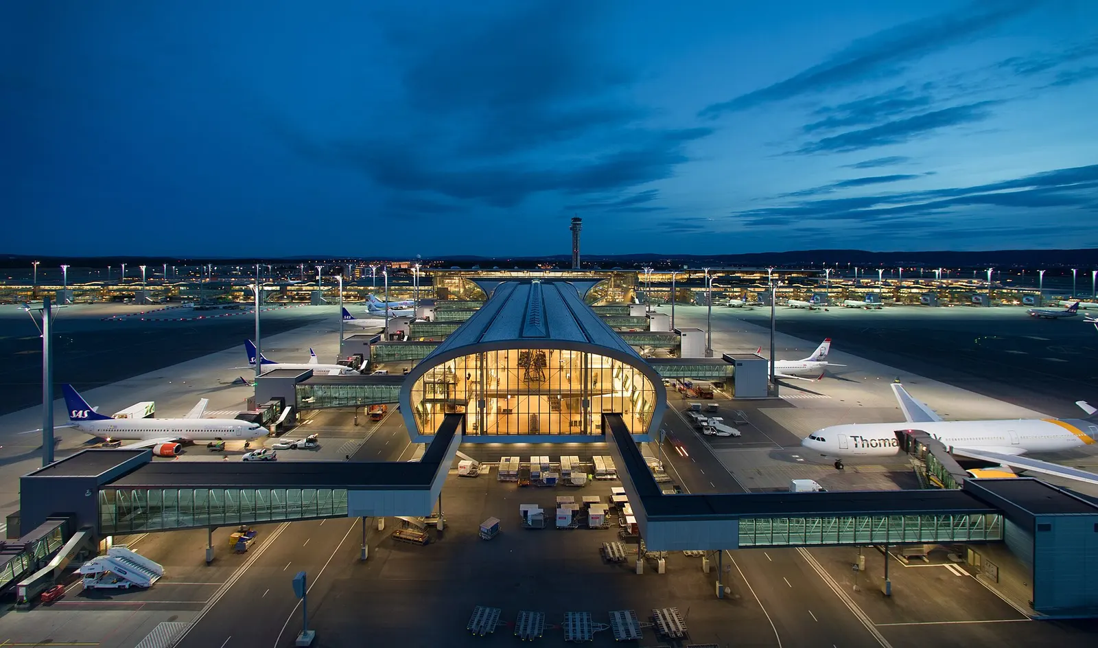 Oslo Airport's illuminated terminal complex at dusk, with warm light glowing through large windows against a deep blue evening sky.