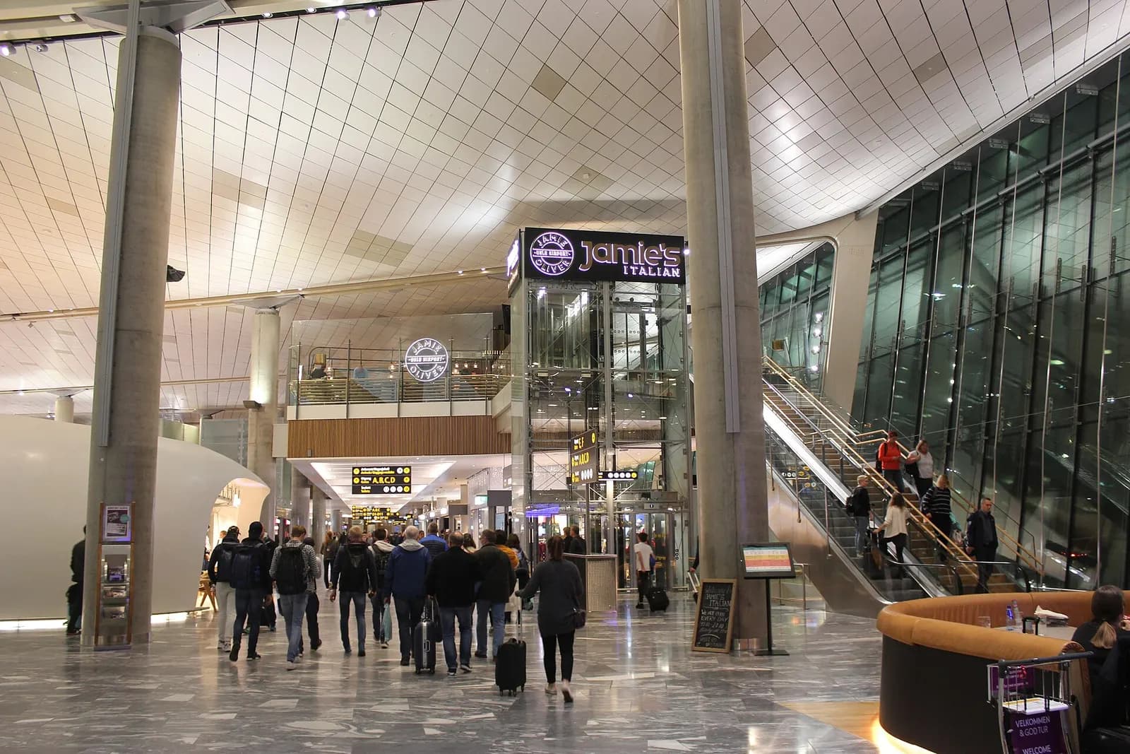 Wide view of Oslo Airport's bright terminal interior with high ceilings, wooden accents, flight information displays, and passengers walking through the main hall.