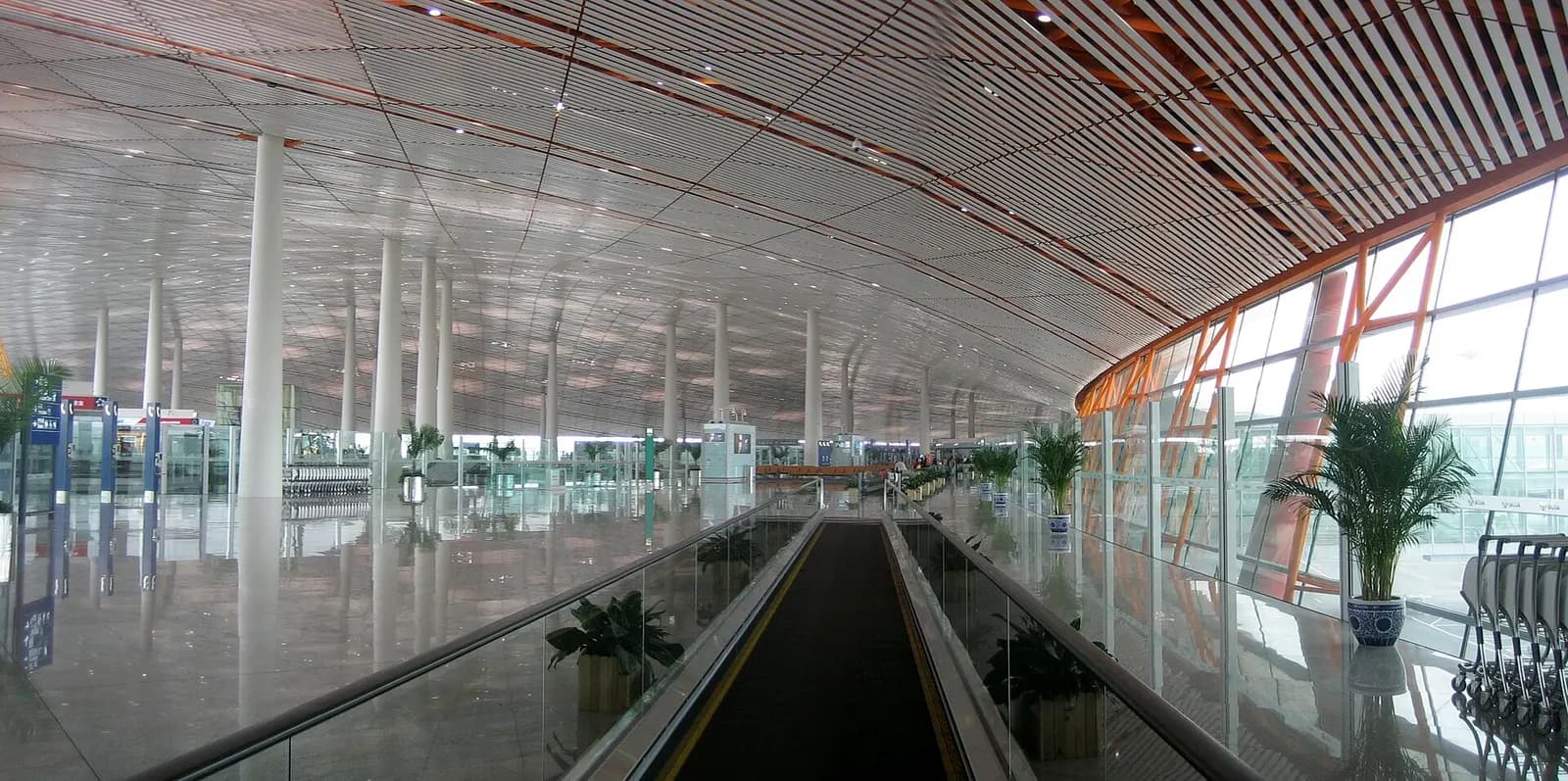 Wide panoramic view of Beijing Capital International Airport Terminal 3, showing its long curved orange roof, glass facade, and drop-off roadway.