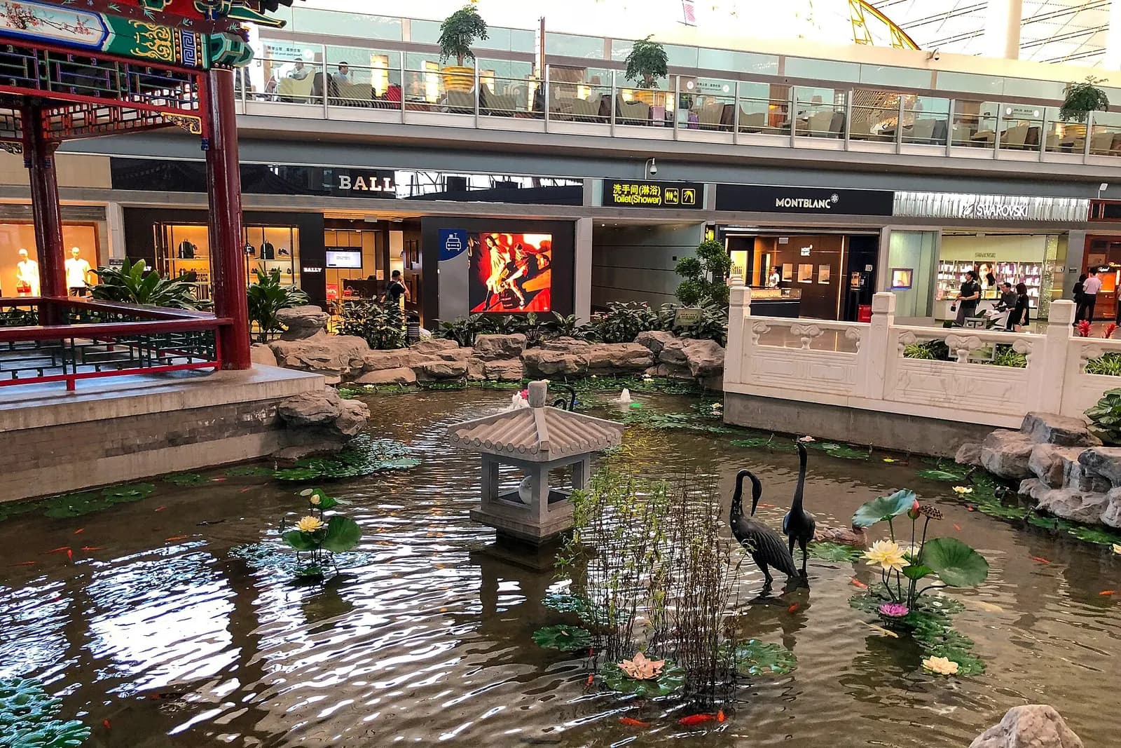 Traditional Chinese garden with a pond, rock formations, and pavilion inside the Terminal 3E international departures concourse.