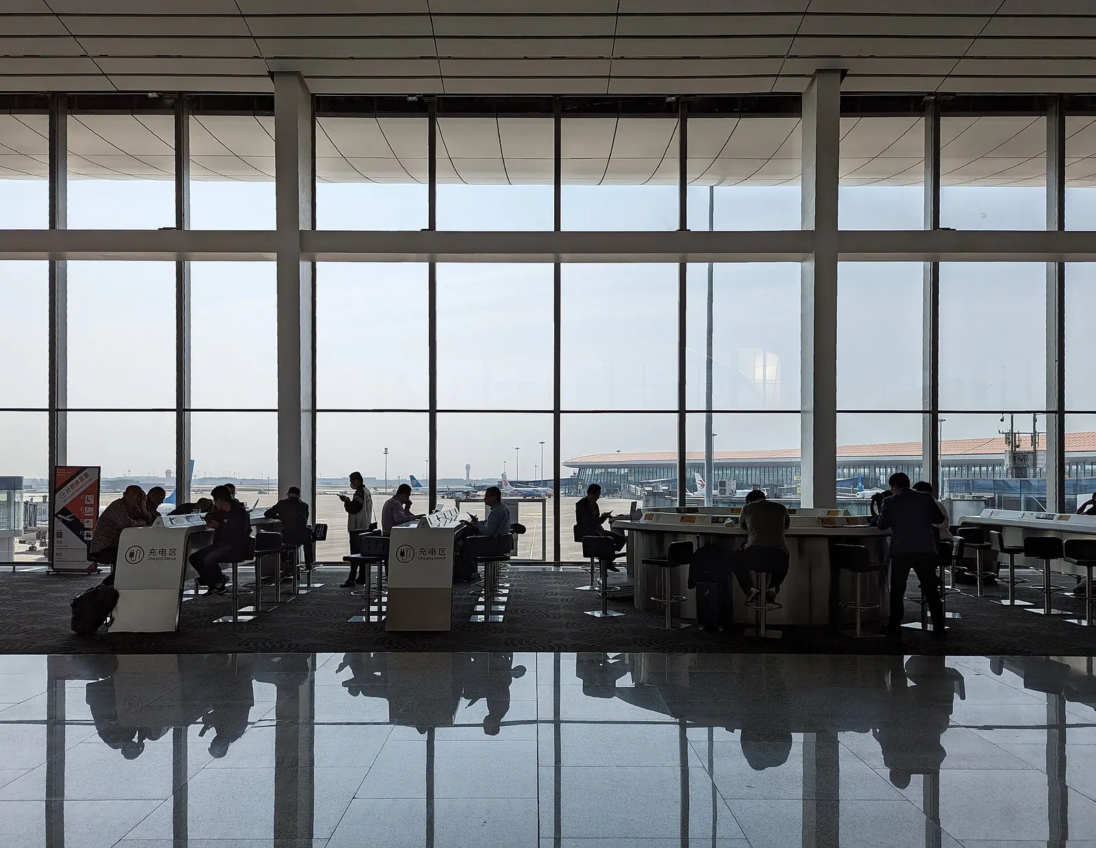 An airside waiting lounge with rows of seats, charging stations, and shopfronts along a wide concourse.