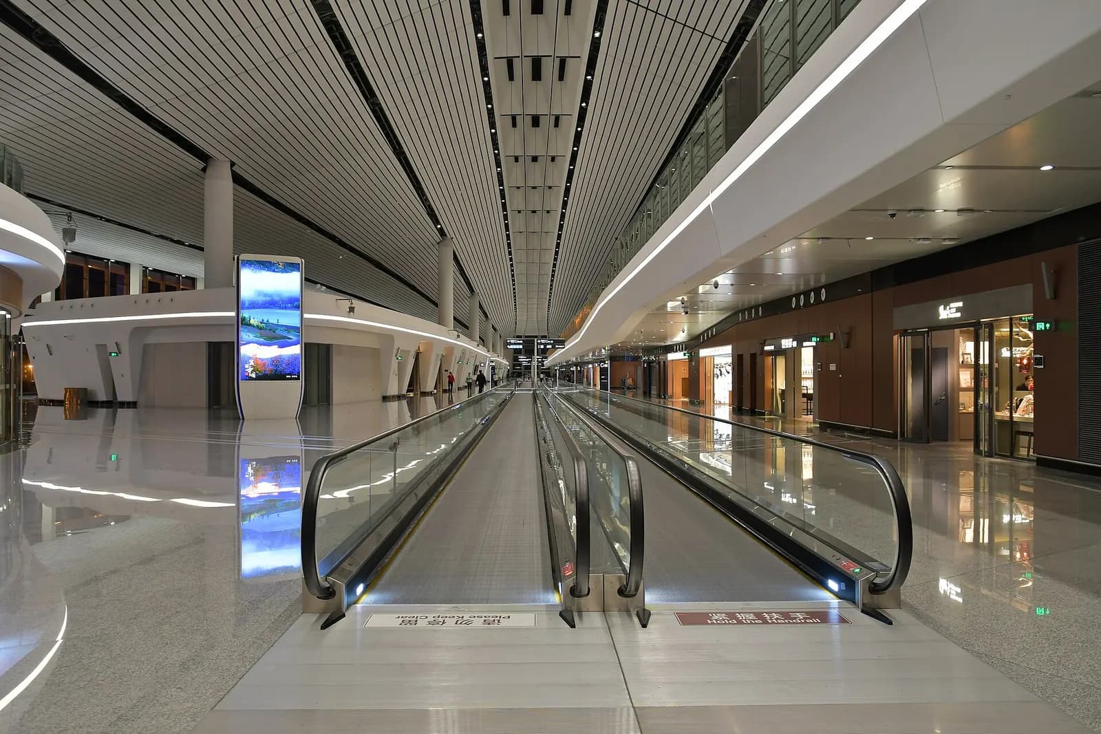 A long, brightly lit concourse corridor with a ribbed white ceiling, moving walkways, and gate signage along the sides.