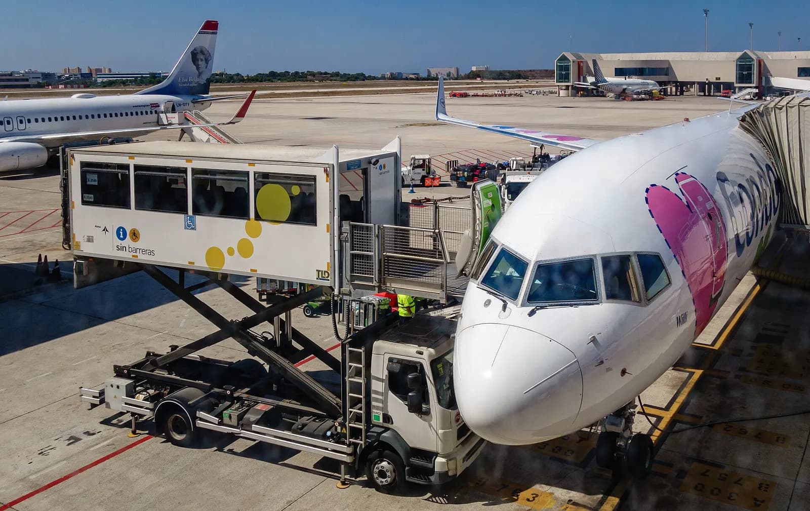 Travelers board a parked aircraft via a jet bridge and ramp stairs, with accessibility equipment set up at the gate.