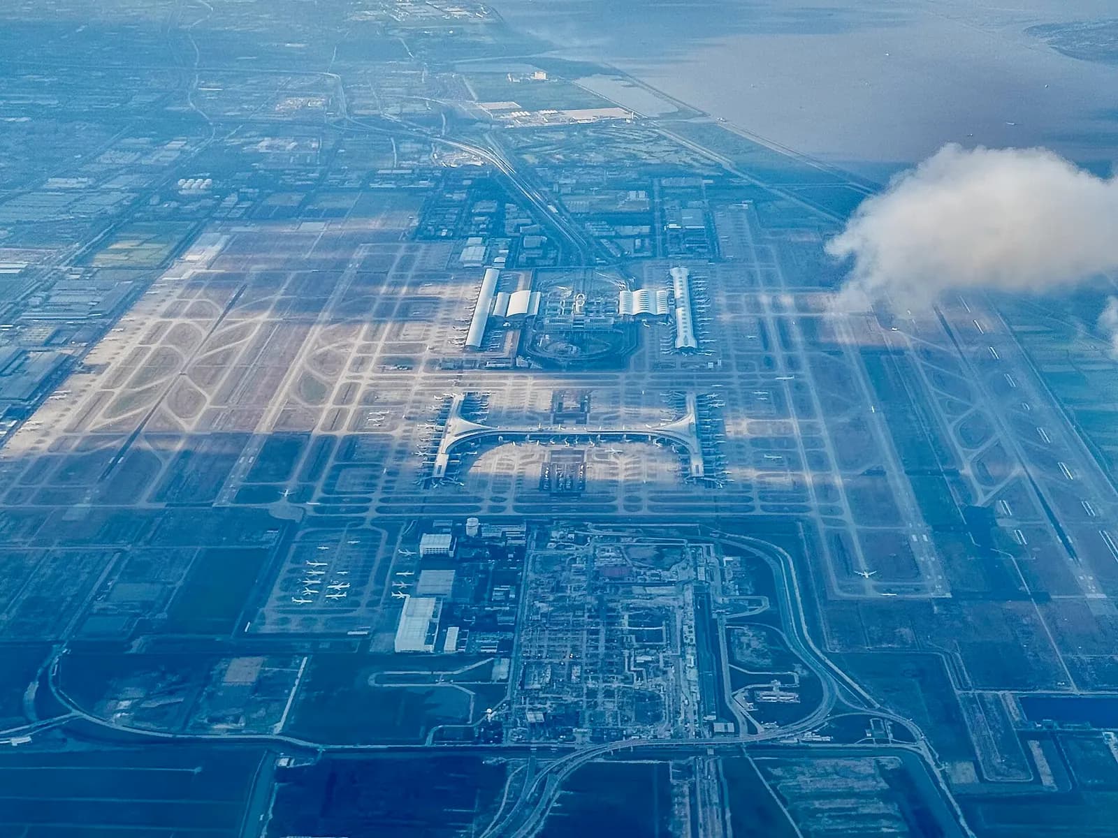 Aerial photograph showing the sprawling layout of Shanghai Pudong International Airport with multiple terminals, runways, and satellite concourses.