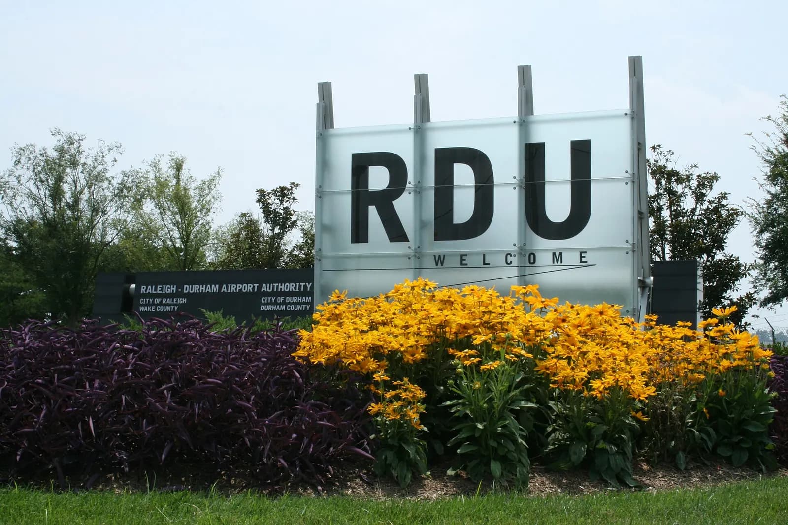 A roadside welcome sign reading Raleigh-Durham International Airport at the Lumley Road entrance, with trees and pavement around it.