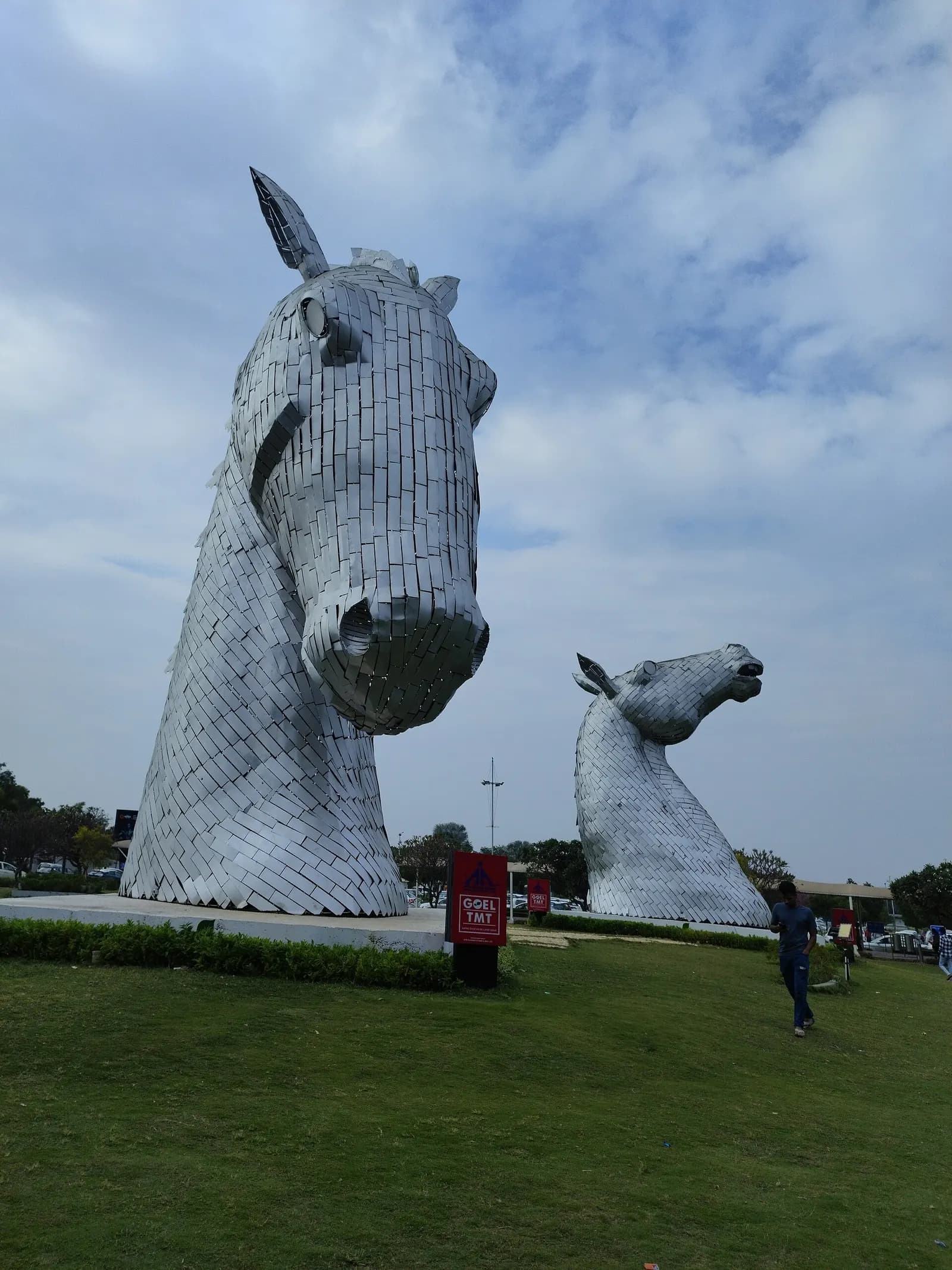 This photograph captures the two monumental horse head sculptures located in the public park area of Swami Vivekananda Airport in Raipur, Chhattisgarh, India. Crafted in a contemporary style, the sculptures are made from hundreds of small, rectangular steel plates welded together. This unique constr