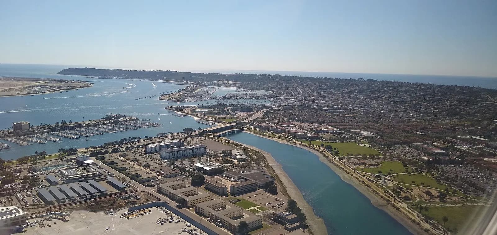 An aerial view of San Diego International Airport's single runway beside San Diego Bay, with Liberty Station park and the city skyline visible nearby.