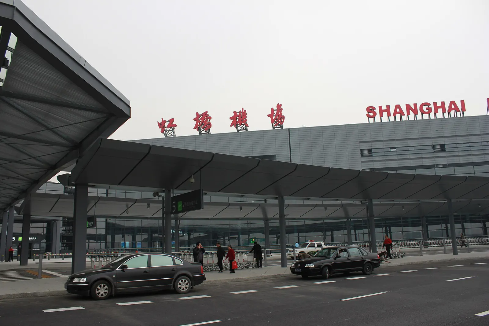 The curved glass-and-steel facade of Shanghai Hongqiao Airport Terminal 2 rises above the frontage roads and passenger drop-off area.