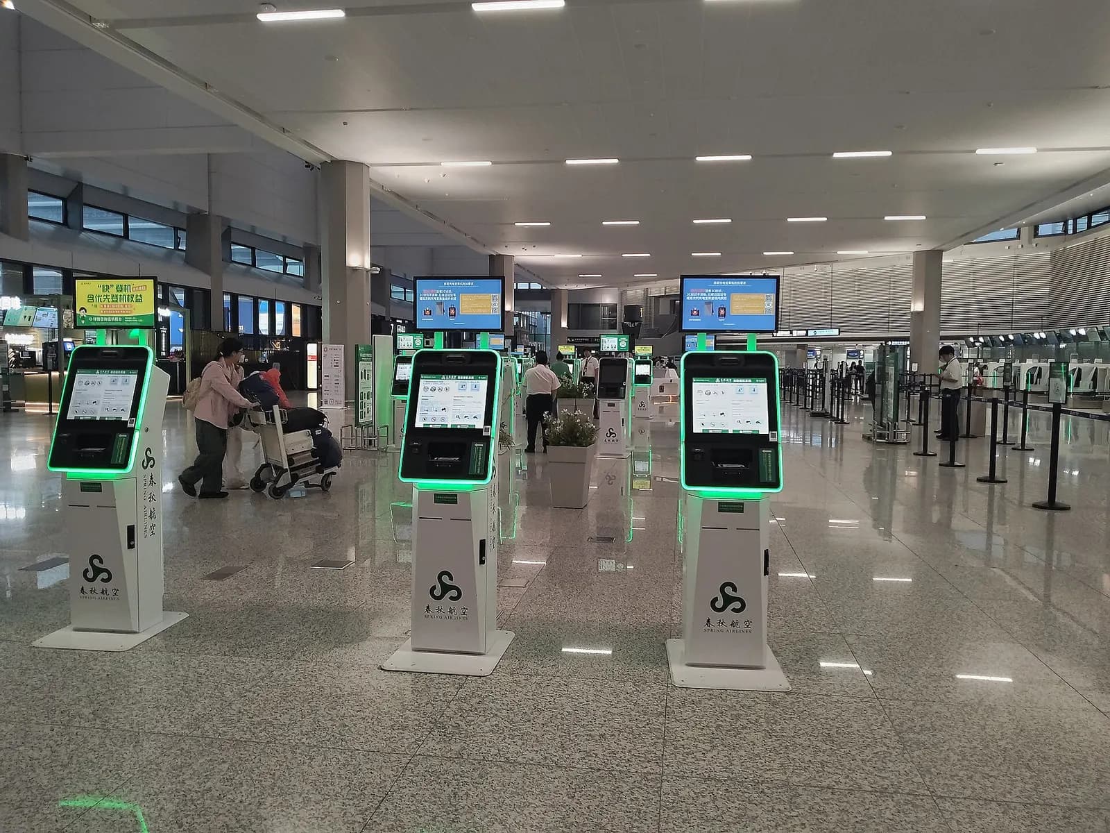 Spring Airlines check-in islands and self-service kiosks line the departure hall of Terminal 1 beneath overhead flight information displays.
