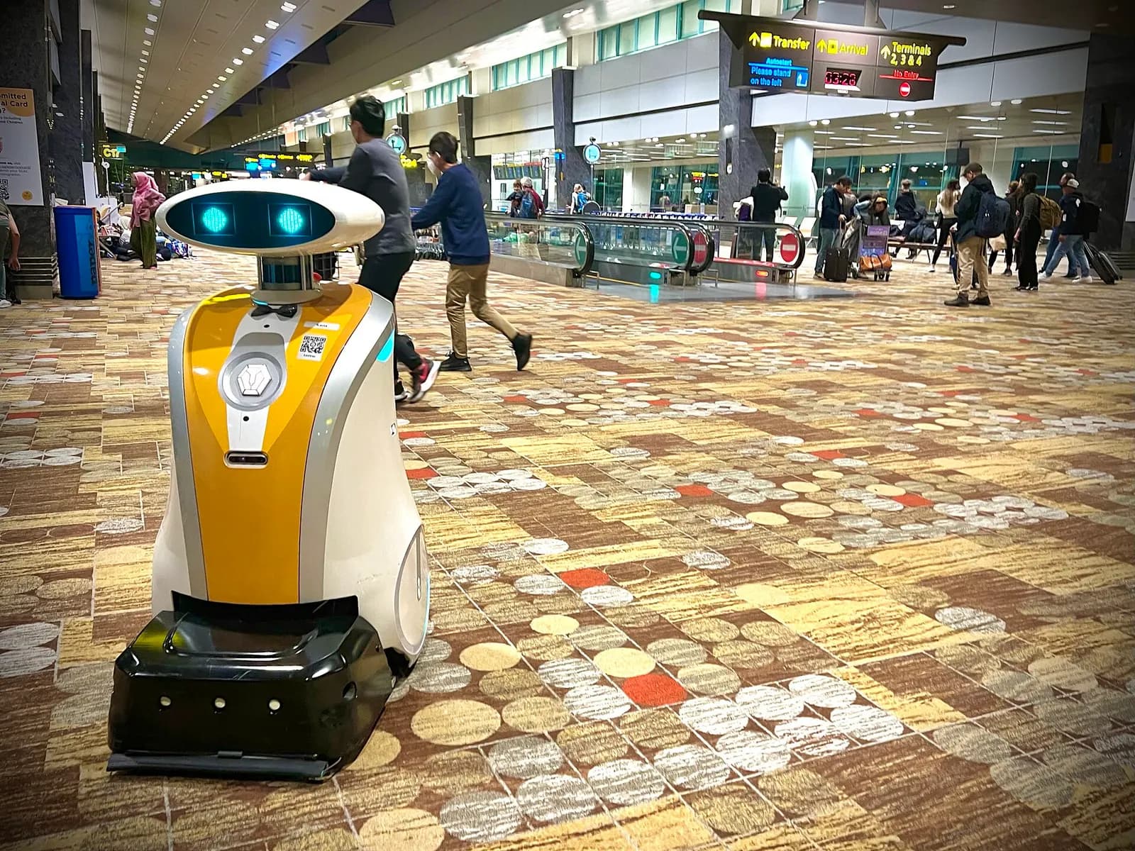 A white service robot on wheels stands in a Changi Airport public area near seating and terminal signage.