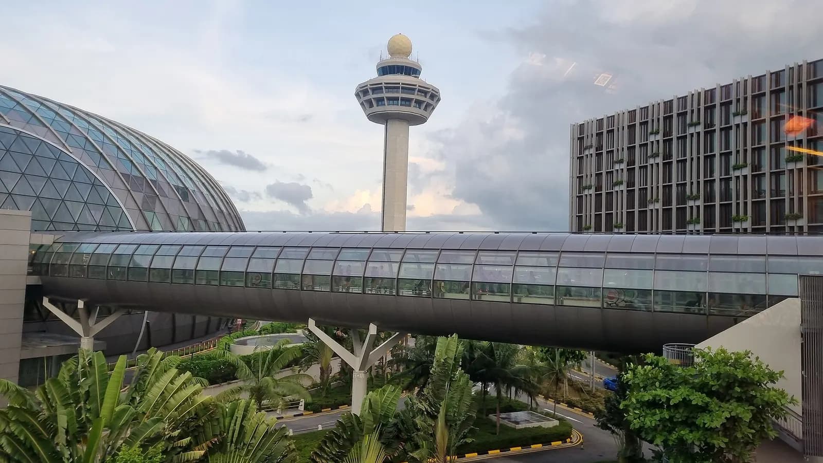 Changi Airport's distinctive control tower rises above terminal buildings connected by an elevated pedestrian walkway under a cloudy sky.