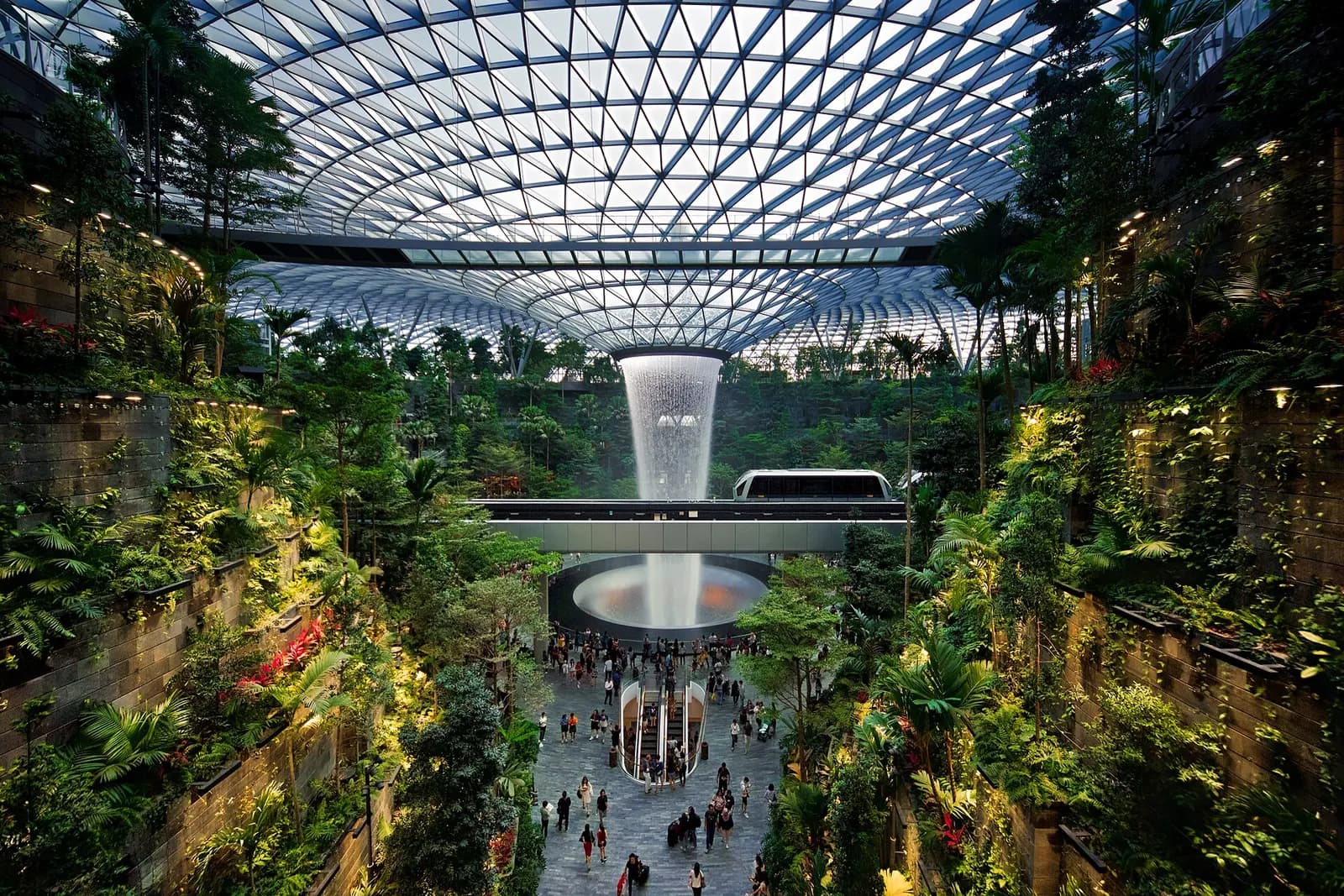 The HSBC Rain Vortex waterfall cascades through the glass-domed atrium of Jewel Changi Airport with lush indoor greenery and an elevated Skytrain track visible above.