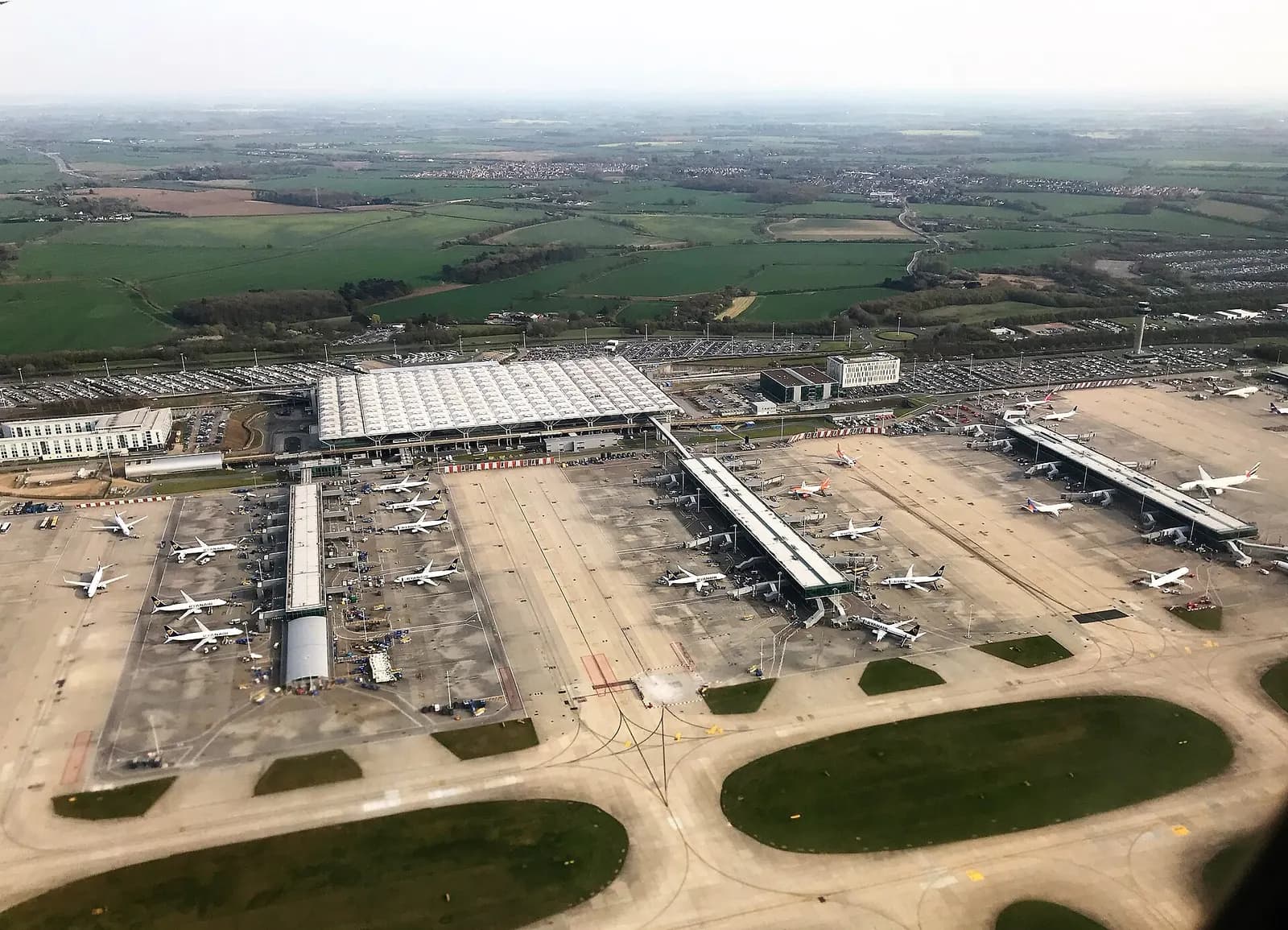 An aerial view of London Stansted Airport showing the runway, terminal, and surrounding Essex countryside.