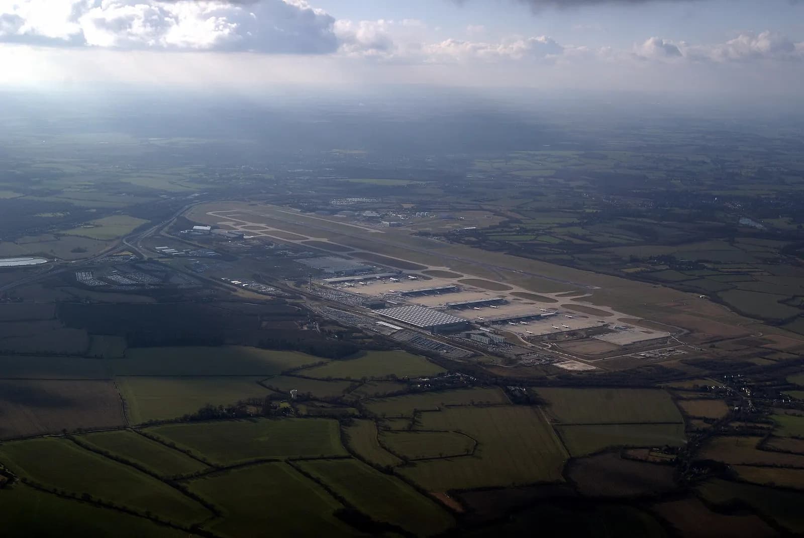 A wide aerial view of the entire London Stansted Airport complex seen from a departing aircraft.