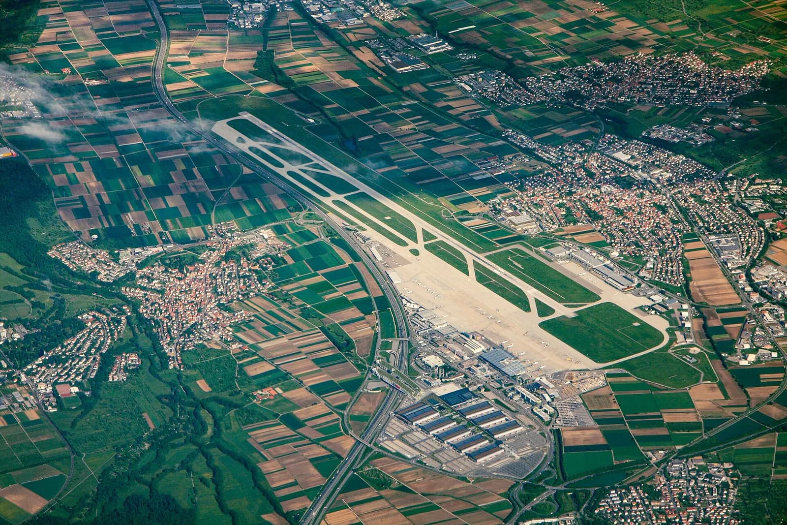 An aerial view of Stuttgart Airport showing parallel runways, terminal buildings, aprons, and surrounding wooded hills.