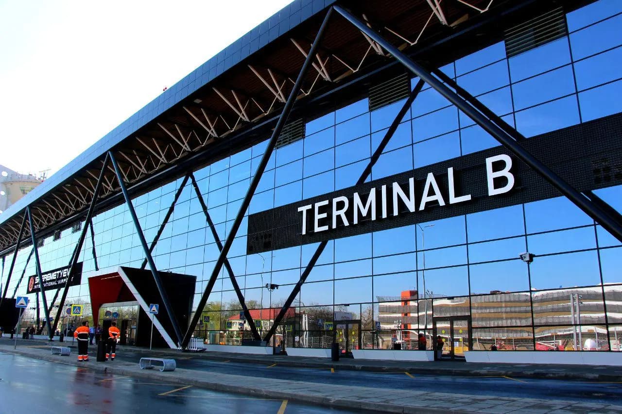 Exterior of Terminal B with a modern glass facade, covered passenger drop-off lanes, and the terminal name visible at the entrance.